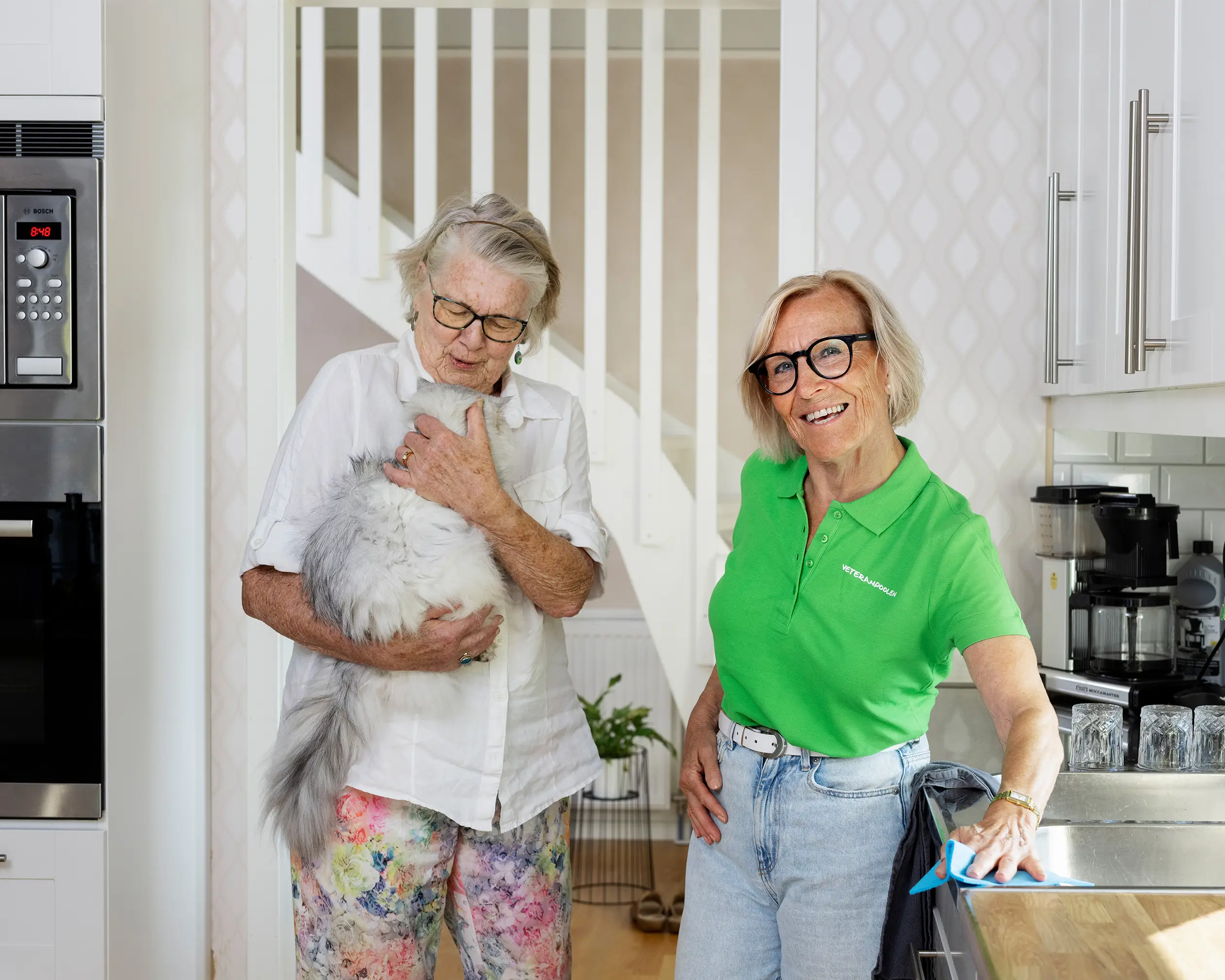 En äldre dam håller i en katt och en kvinna står bredvid, de står i ett kök