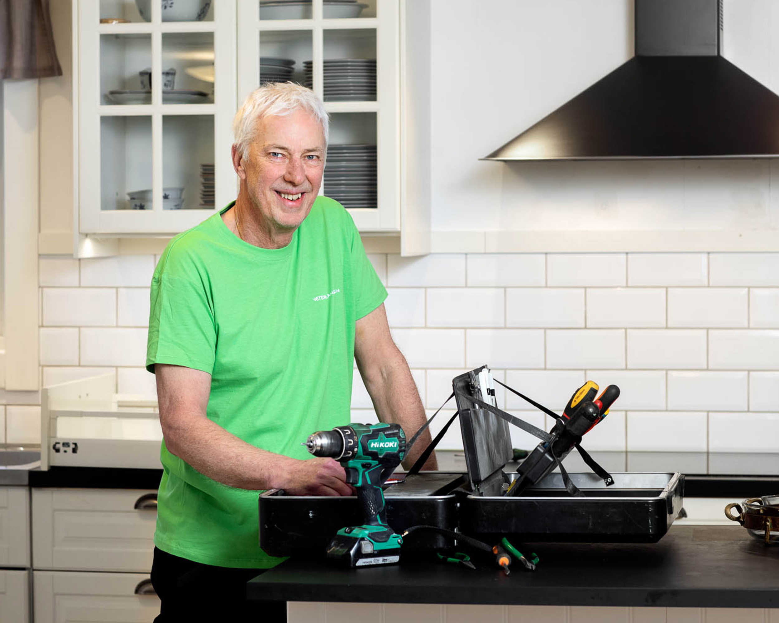 En veteran har en verktygslåda och står i ett kök En veteran står i ett kök och har en verktygslåda, han har en grön t-shirt på sig och ler mot kameran