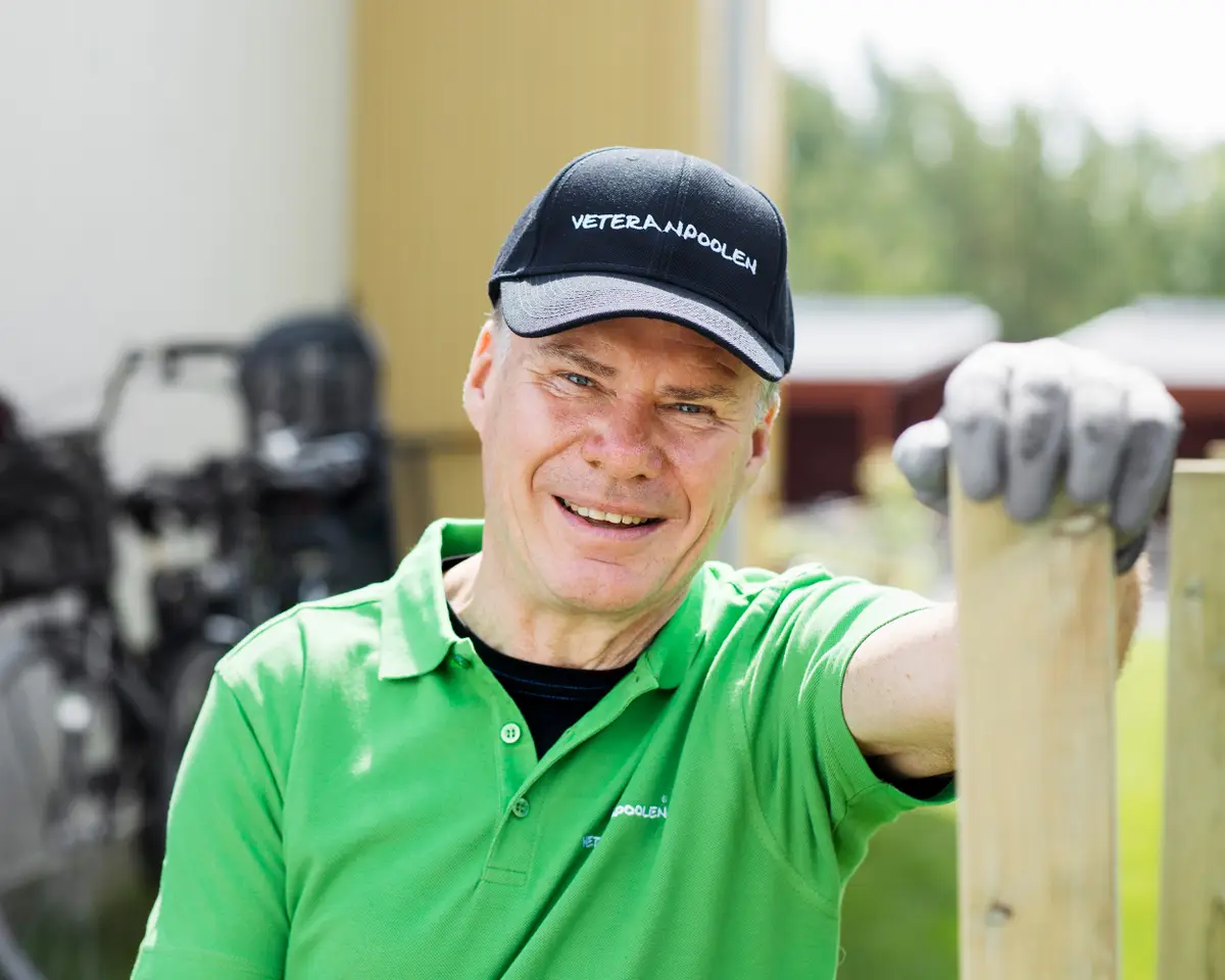 En man står och håller handen på en staketstolpe, han har en grön tröja på sig och ler mot kameran