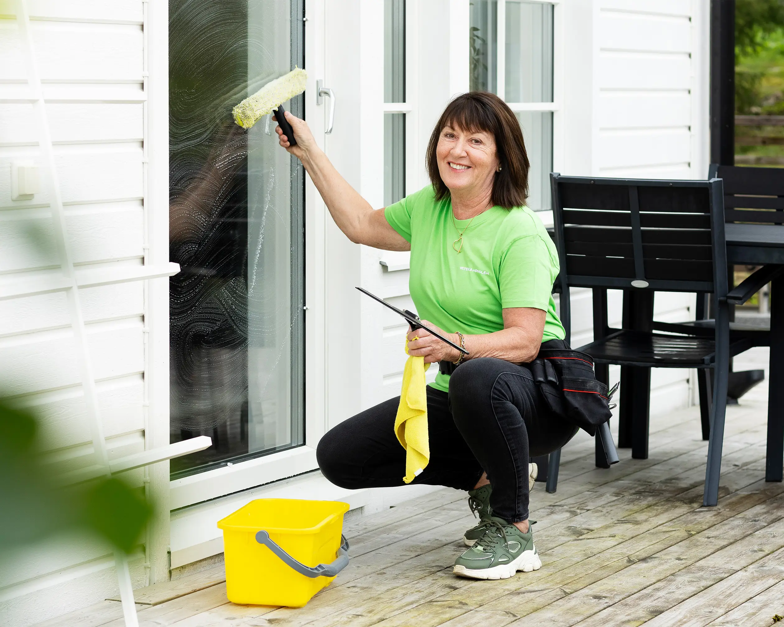 Pensionerad kvinna i grön t-shirt putsar fönster och ler mot kameran i dagsljus.