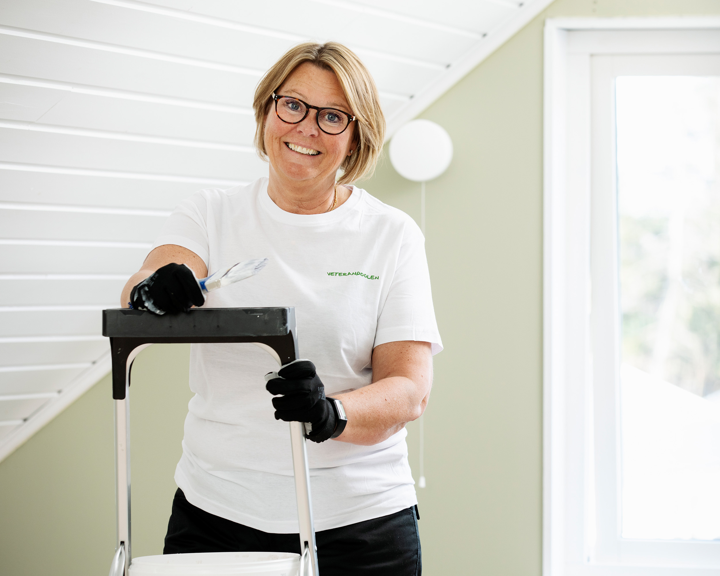 En kvinnlig hantverkare står med målarpensel i handen En kvinnlig hantverkare står med målarpensel i handen, hon ler mot kameran och ska måla ett innertak.