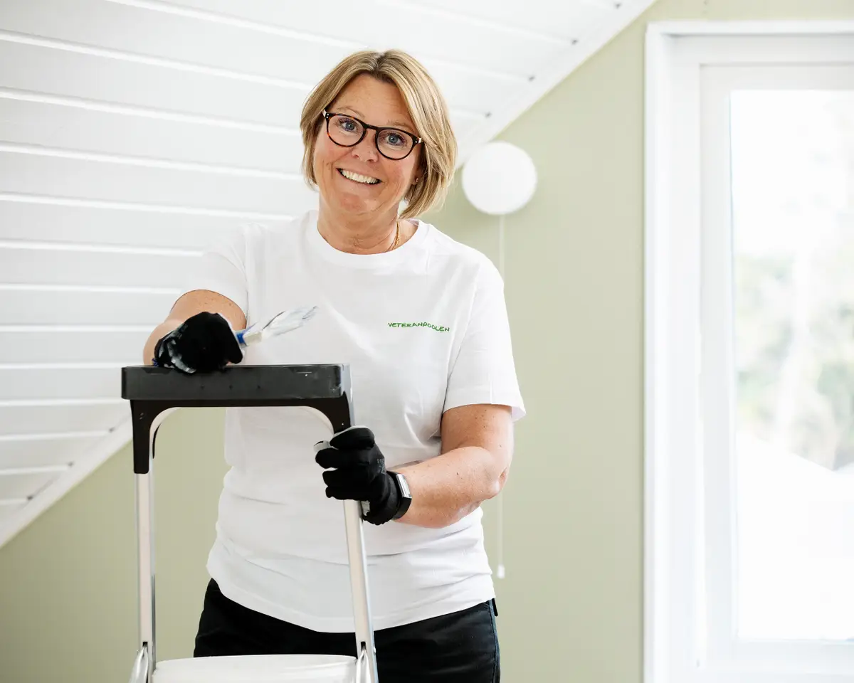 En kvinnlig hantverkare står med målarpensel i handen, hon ler mot kameran och ska måla ett innertak.