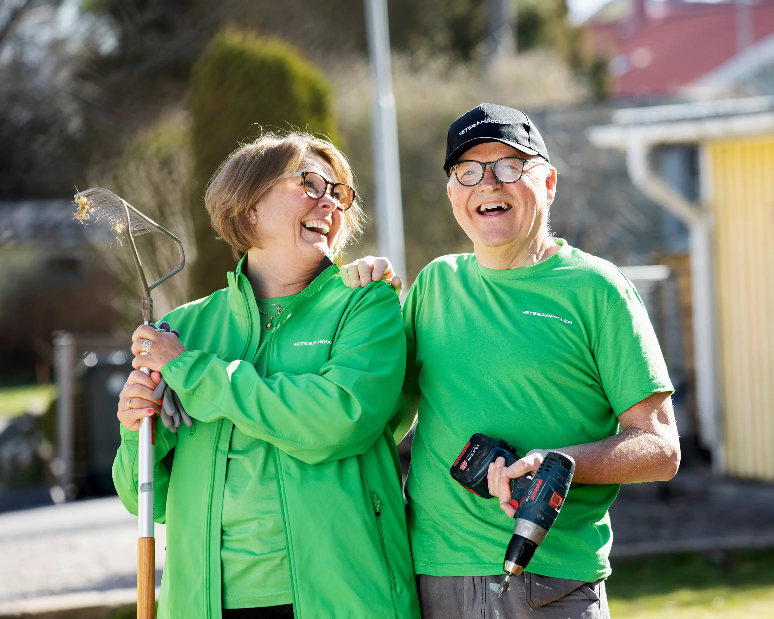 Två jobbonärer står i gröna tröjor ute i en trädgård, de ler och mannen tittar mot kameran