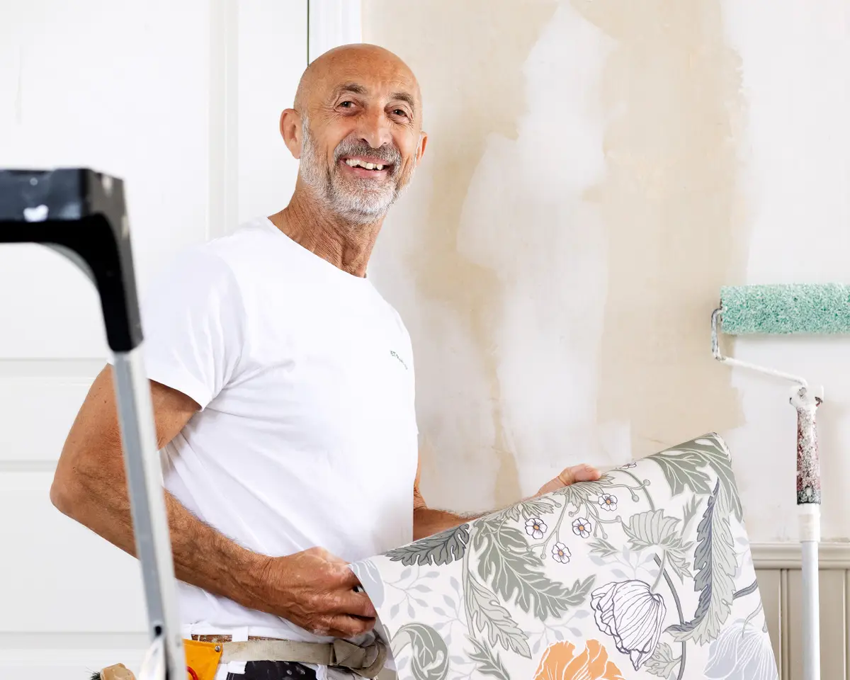 En målare håller i en tapet, han ska tapetsera en vägg bakom honom