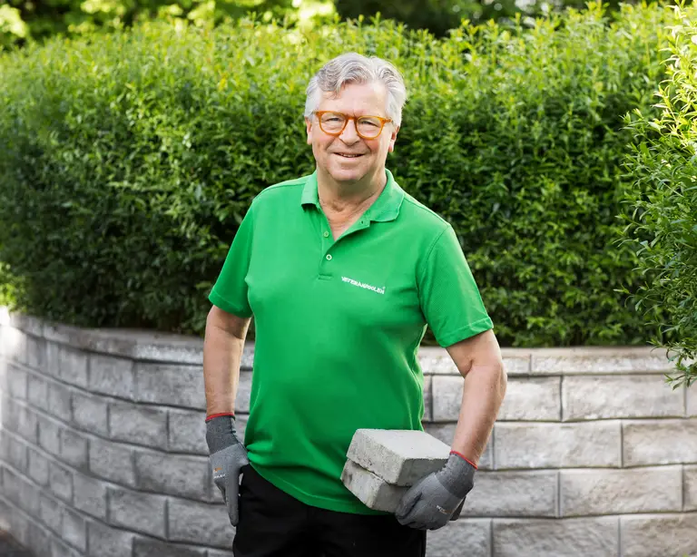 En hantverkare står med två murblock i famnen, han står framför en mur