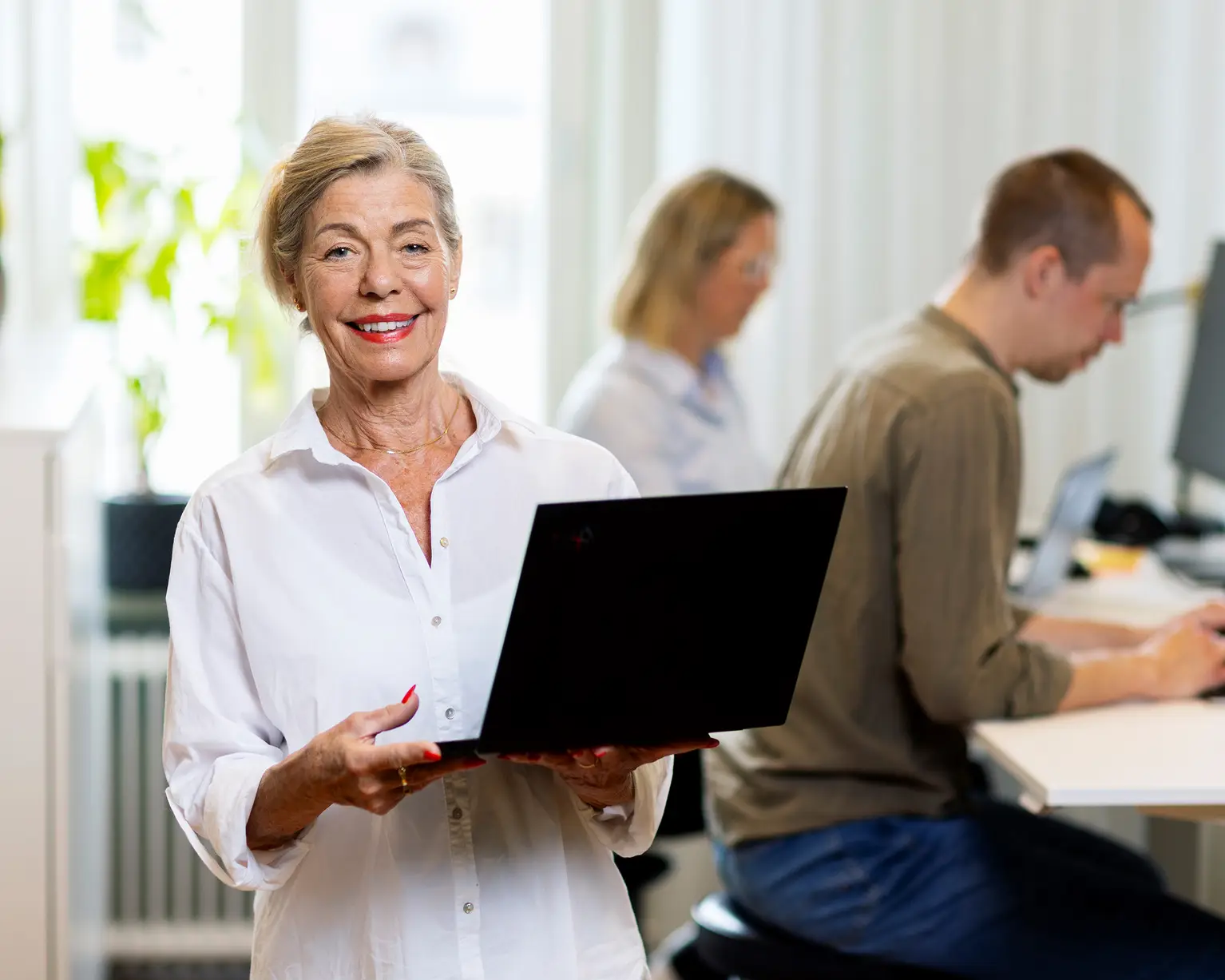 En kvinnlig jobbonär står med en laptop i famnen och tittar mot kameran, hon ler och har på sig en vit skjorta.
