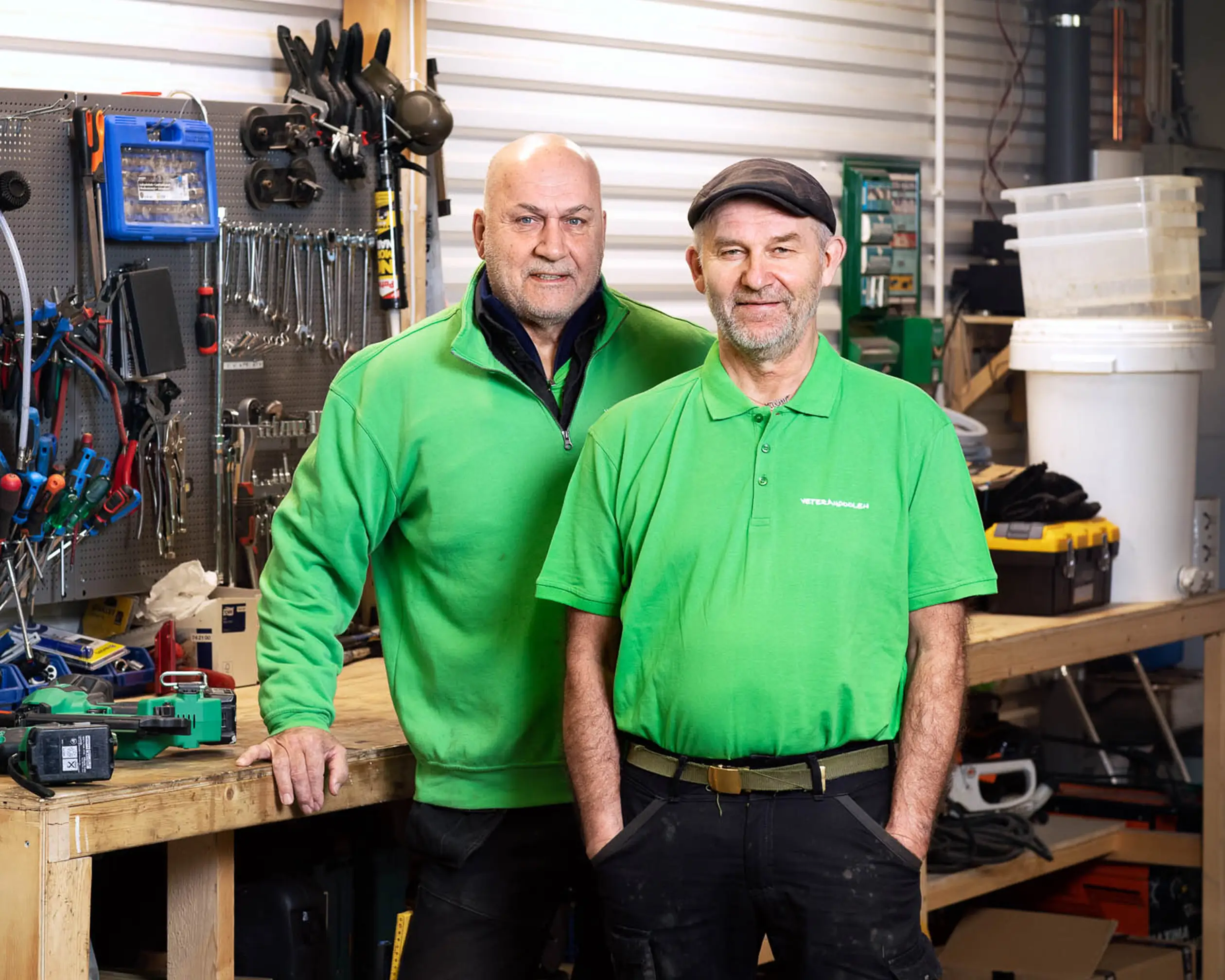 Två manliga jobbonärer med gröna tröjor står i ett förråd med verktyg