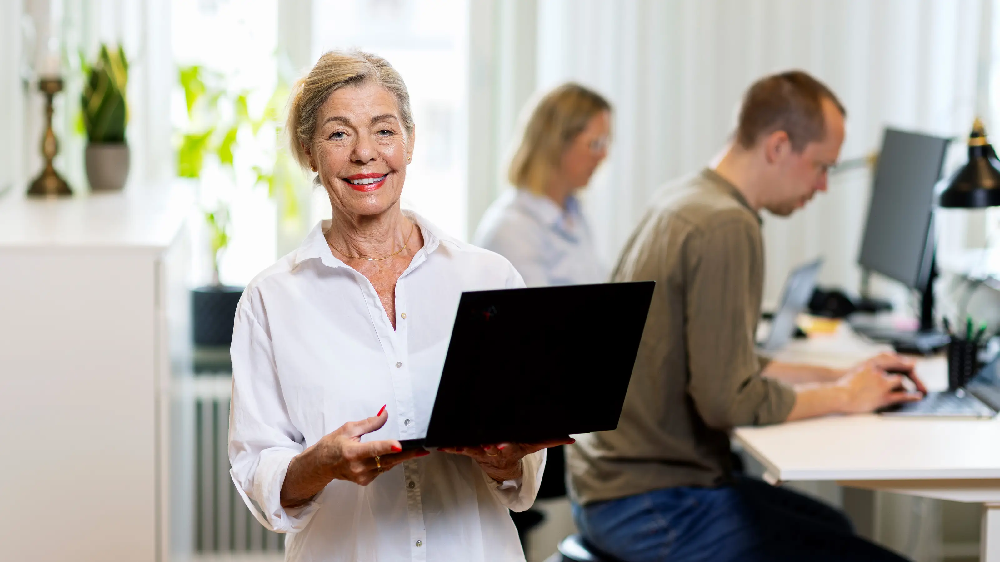 En kvinnlig jobbonär står med en laptop i famnen och tittar mot kameran, hon ler och har på sig en vit skjorta.