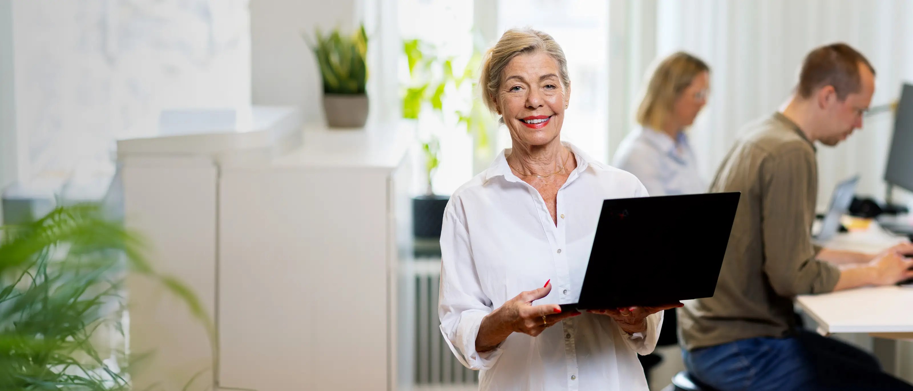 En kvinnlig jobbonär står med en laptop i famnen och tittar mot kameran, hon ler och har på sig en vit skjorta.