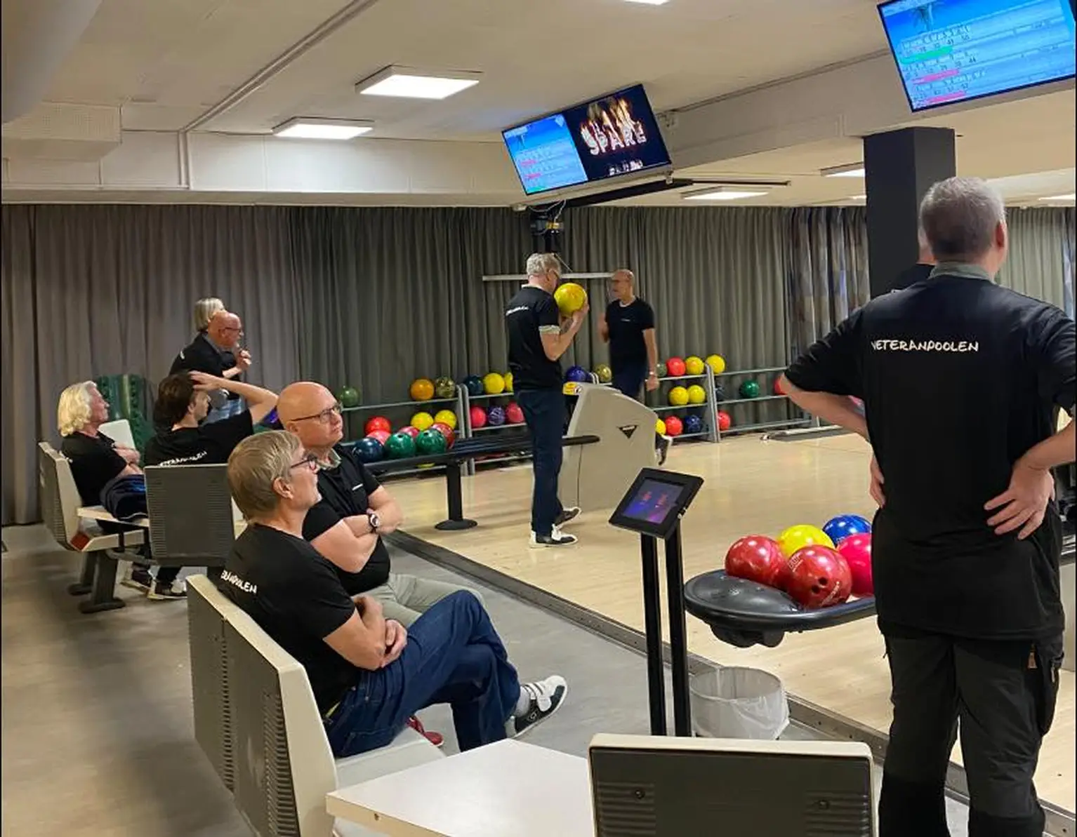 People in black shirts are gathered in a bowling alley, some sitting, others preparing to bowl, with colorful balls visible nearby.