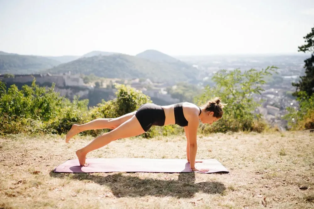 Person practicing yoga outdoors on a mat, balancing on hands and one leg. Scenic view of hills and greenery in the background.