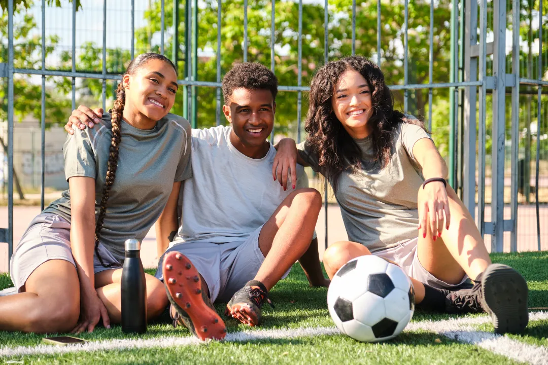3 Personen sitzen am Fußballplatz am Boden mit einem Ball und lächeln