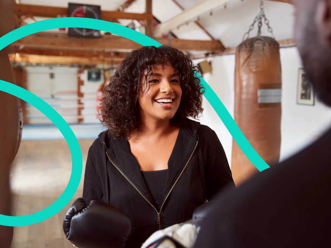 Eine Frau in einem Fitnessstudio lächelt, während sie Boxhandschuhe hält, im Hintergrund sind Boxsäcke zu sehen.