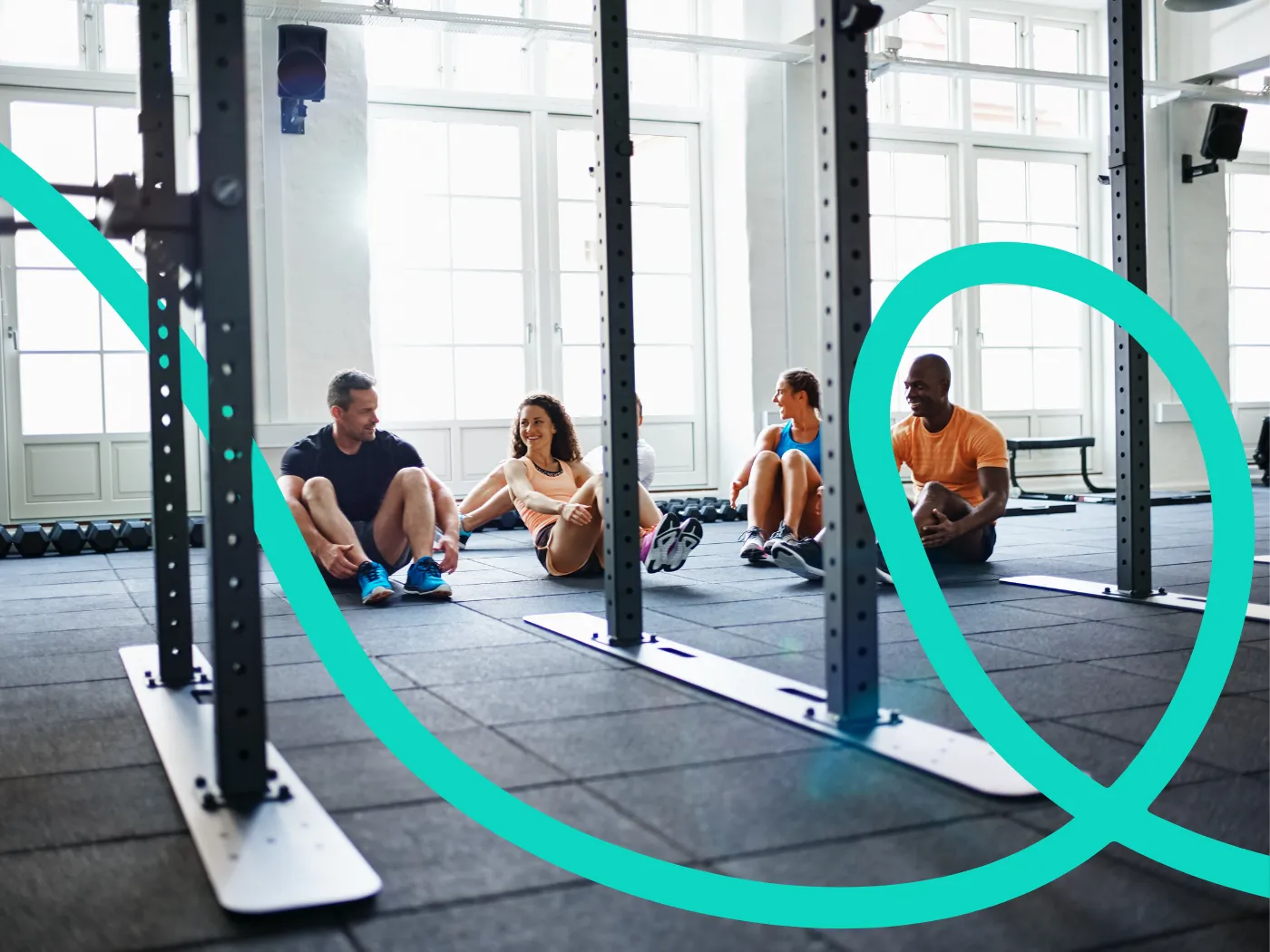 Vier Personen sitzen auf dem Boden eines Fitnessstudios, lächeln und unterhalten sich, im Hintergrund sind Trainingsgeräte und große Fenster zu sehen.