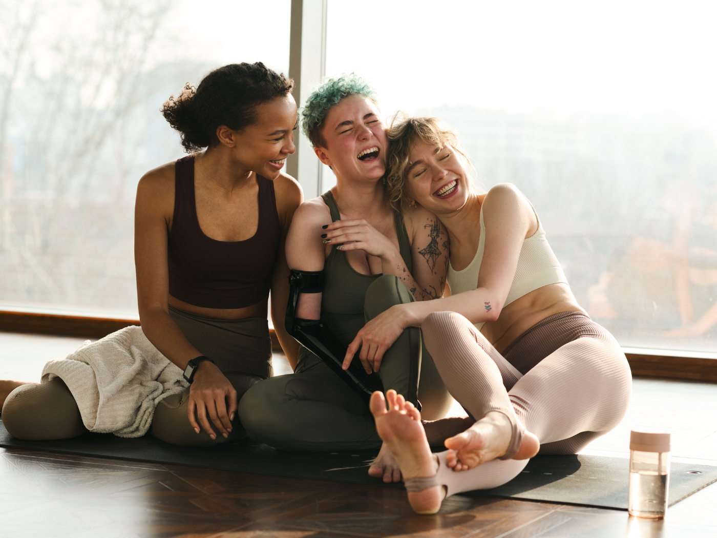 3 women sitting on the floor in fitness clothing on a yoga mat and laughing