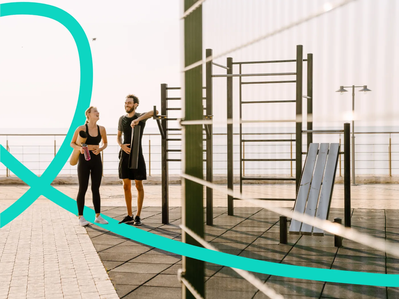 Zwei Personen in Sportkleidung stehen am Meer bei Outdoor-Trainingsgeräten, unterhalten sich und halten eine Wasserflasche.