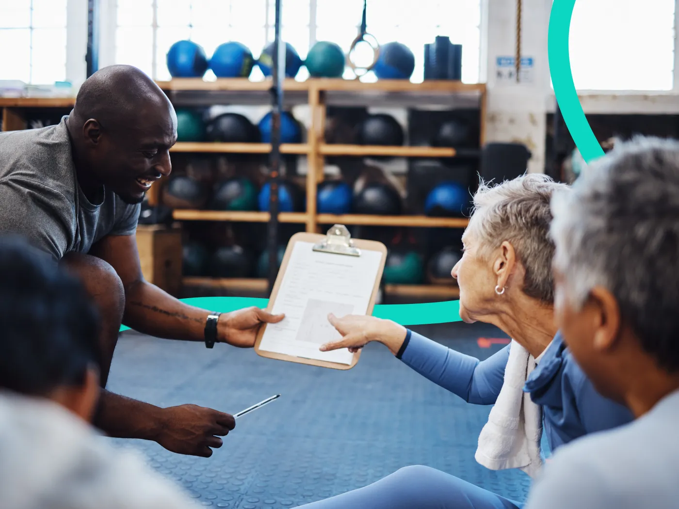 Ein Trainer zeigt zwei sitzenden älteren Erwachsenen in einem Fitnessstudio ein Klemmbrett, umgeben von Trainingsgeräten und Medizinbällen.