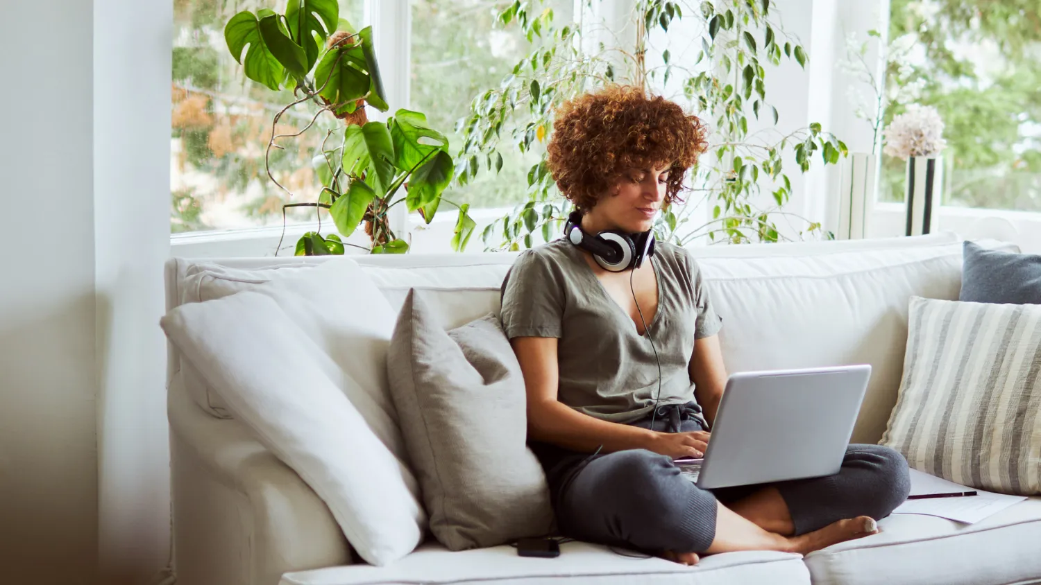 Person with curly hair sits cross-legged on a white couch, wearing headphones and using a laptop, surrounded by plants and large windows.