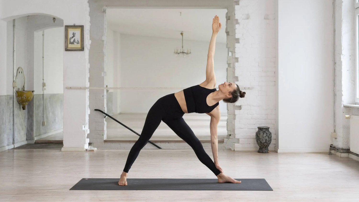 Woman practicing yoga in a bright room, performing a triangle pose on a black mat with one arm extended upward.