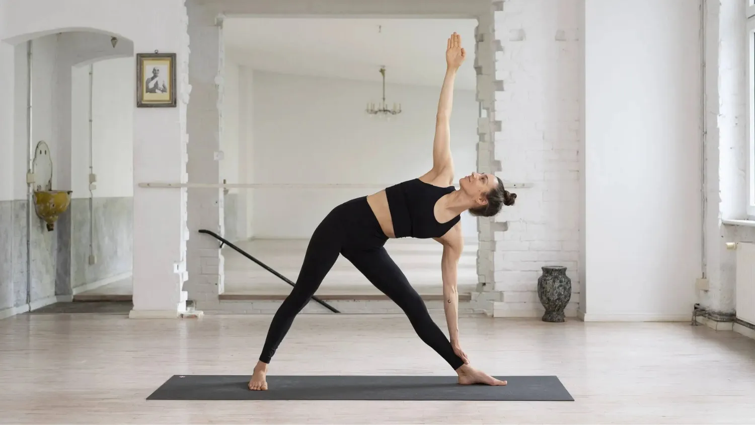 Woman practicing yoga in a bright room, performing a triangle pose on a black mat with one arm extended upward.