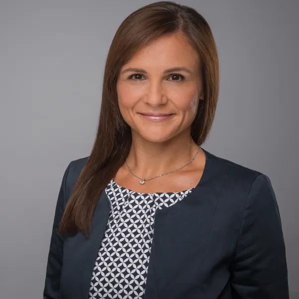 Woman with long brown hair in a dark blazer and patterned blouse, smiling against a gray background.