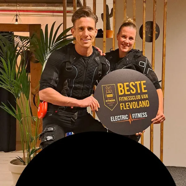 Two people smiling and holding a sign that reads "Beste Fitnessclub van Flevoland, Electric Fitness," with plants in the background.