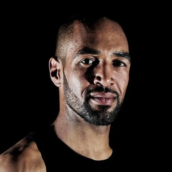 Man with a beard and short hair, wearing a sleeveless black shirt, against a dark background.
