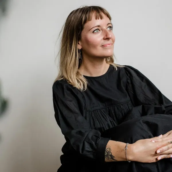 Woman with a tattoo on her forearm, wearing a black dress, sitting and looking to the side against a plain background.