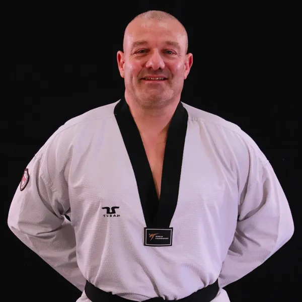 A person wearing a white martial arts uniform with a black belt stands against a black background, smiling with hands on hips.