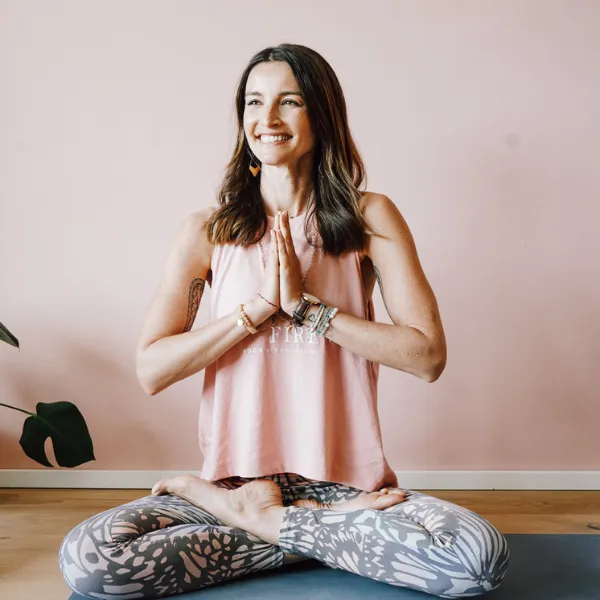Woman smiling in a seated yoga pose, wearing a pink top and patterned leggings, against a soft pink wall.