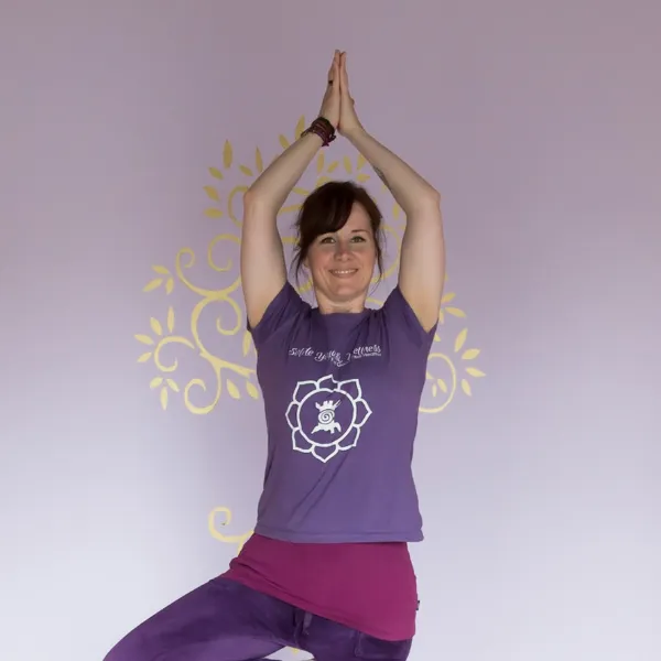 A woman in a purple outfit practices yoga, standing in a tree pose against a light purple wall with a decorative design.