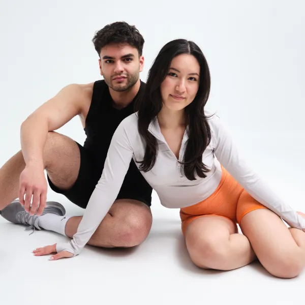 A man and woman sit on the floor in athletic wear. The man wears a black tank top and shorts; the woman wears a white top and orange shorts.
