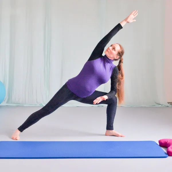 Pregnant woman practicing yoga in a bright room, performing a side angle pose on a blue mat, wearing a purple top and black leggings.