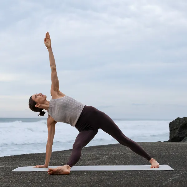 A person practices yoga on a mat by the ocean, performing a side angle pose under a cloudy sky.