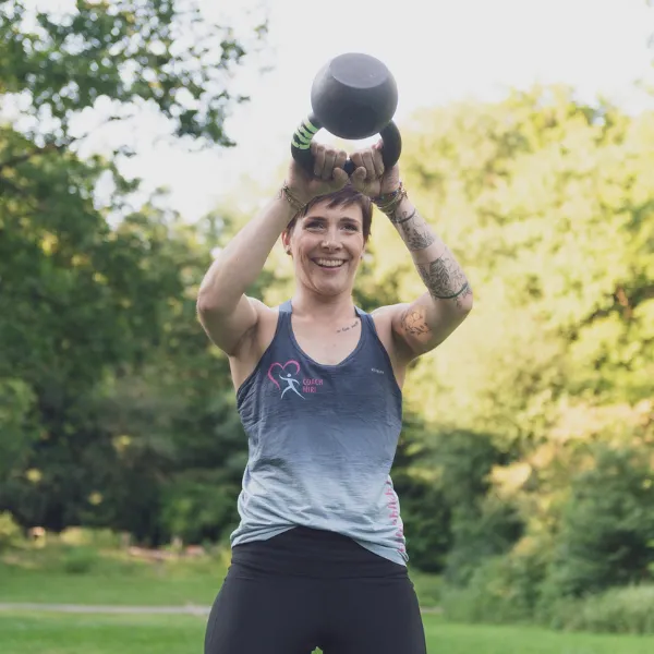 Woman smiling while lifting a kettlebell outdoors, surrounded by greenery. She wears a sleeveless top and has tattoos on her arms.