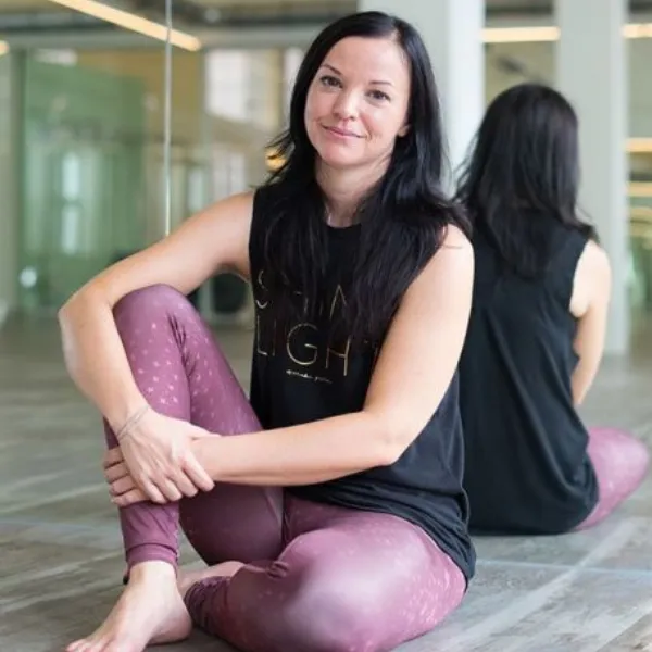 A woman with long dark hair sits on a yoga studio floor, wearing a black tank top and pink leggings, smiling softly with her reflection in the mirror.