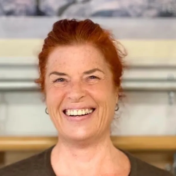 Smiling person with red hair and a brown top, standing indoors against a blurred background.