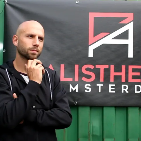 Bald man in a black hoodie stands pensively with hand on chin in front of a sign reading "ALISTHE AMSTERDAM" on a green background.
