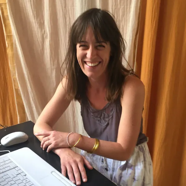 Smiling woman with long hair sits at a desk with a keyboard and mouse, wearing a sleeveless top and skirt, against a curtain backdrop.