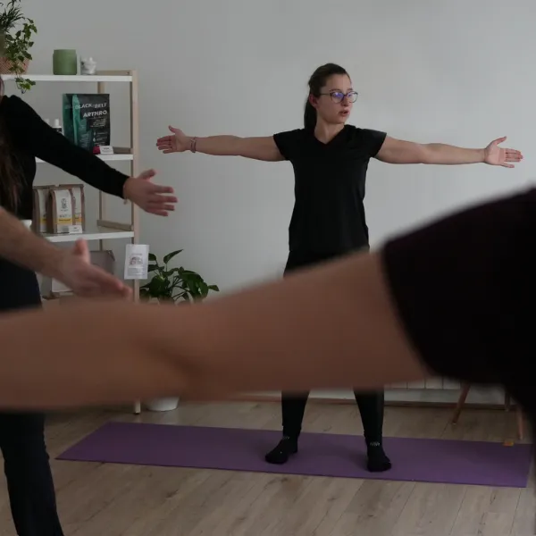 A woman in a black outfit practices yoga on a purple mat, arms outstretched. Others join in the session in a softly lit room with plants.