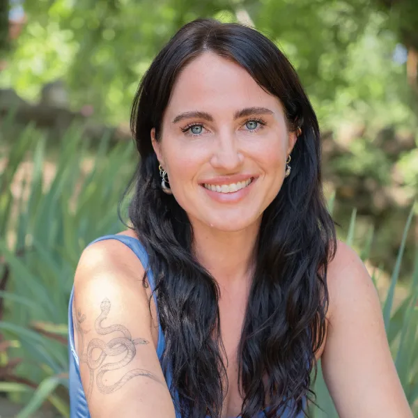 Smiling person with long dark hair, wearing a blue top, sits outdoors with greenery in the background. A snake tattoo is visible on their arm.