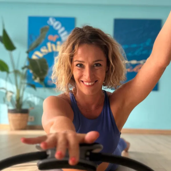 Woman in a blue top smiling and stretching with a fitness ring in a brightly lit room with plants and colorful wall art.