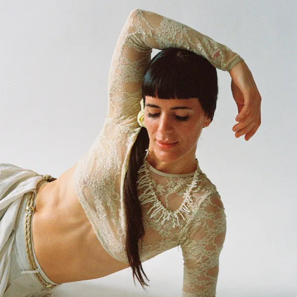 Person in a lace top poses gracefully against a white background, with one arm raised and eyes closed, wearing a beaded necklace.