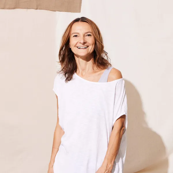 Smiling woman with shoulder-length brown hair wearing a white top stands against a neutral background.
