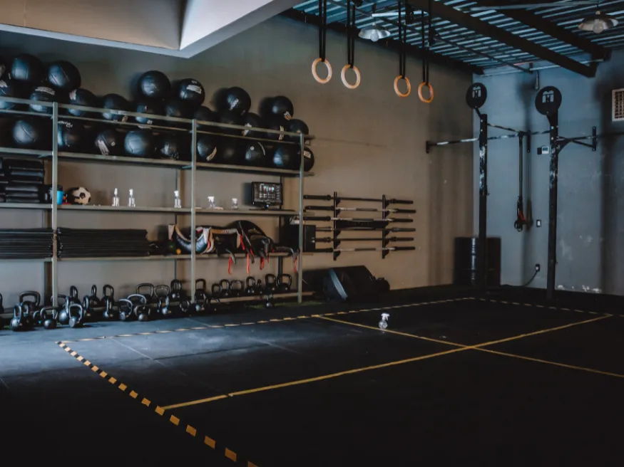 Leeres Fitnessstudio mit Kettlebells, Medizinbällen und Turnringen. Schwarzer Boden mit gelben Linien und verschiedenem Trainingsgerät auf Regalen.