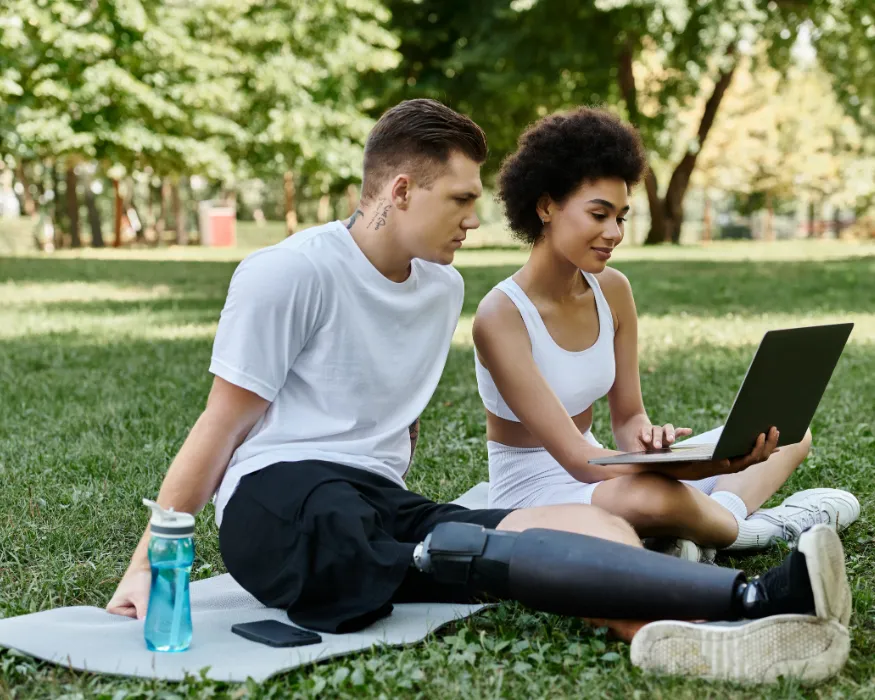 Zwei Personen in Sportkleidung sitzen im Freien auf einer Matte und verwalten ihre Online-Zahlungen am Laptop.