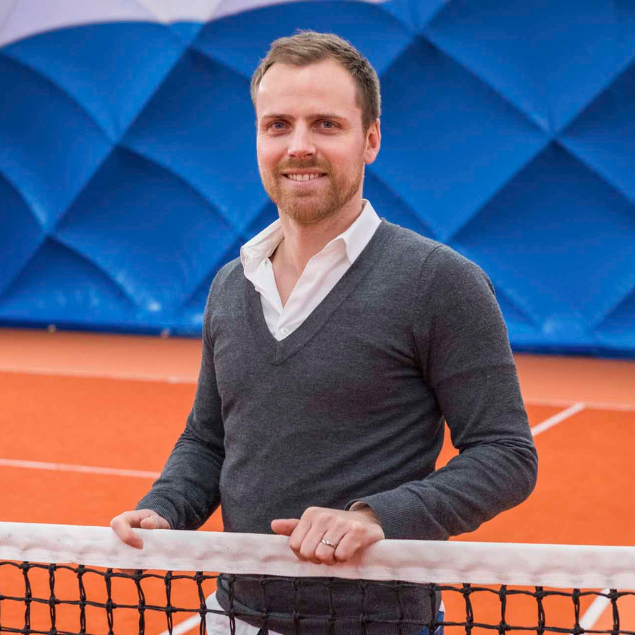Man in a gray sweater and white shirt smiling while leaning on a tennis net, with a blue geometric pattern background.