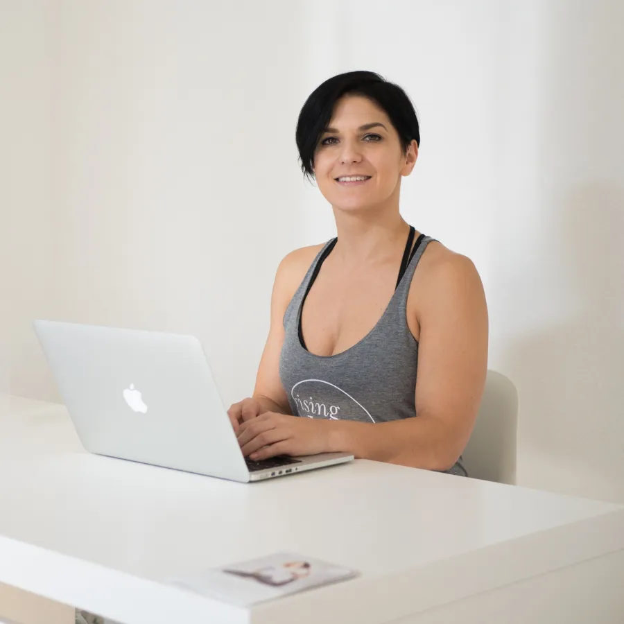 Woman with short dark hair, wearing a gray tank top, sitting at a white desk using a laptop, smiling at the camera.