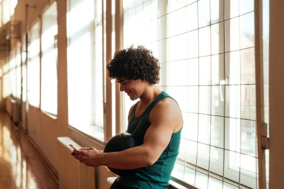 Man with curly hair in a green tank top leans against a wire mesh, holding a ball and looking at his phone, in a sunlit hallway.