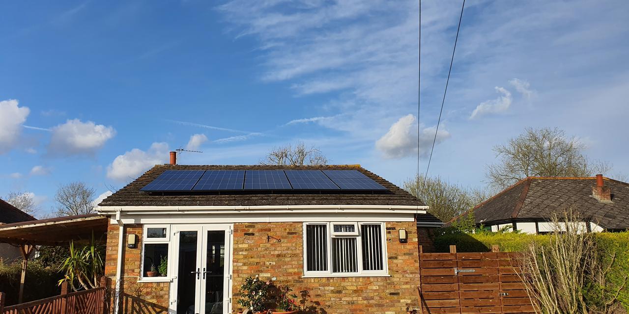 Single-storey brick house with solar panels on the roof, set under a bright blue sky with scattered clouds, surrounded by neighbouring homes, plants and a wooden fence